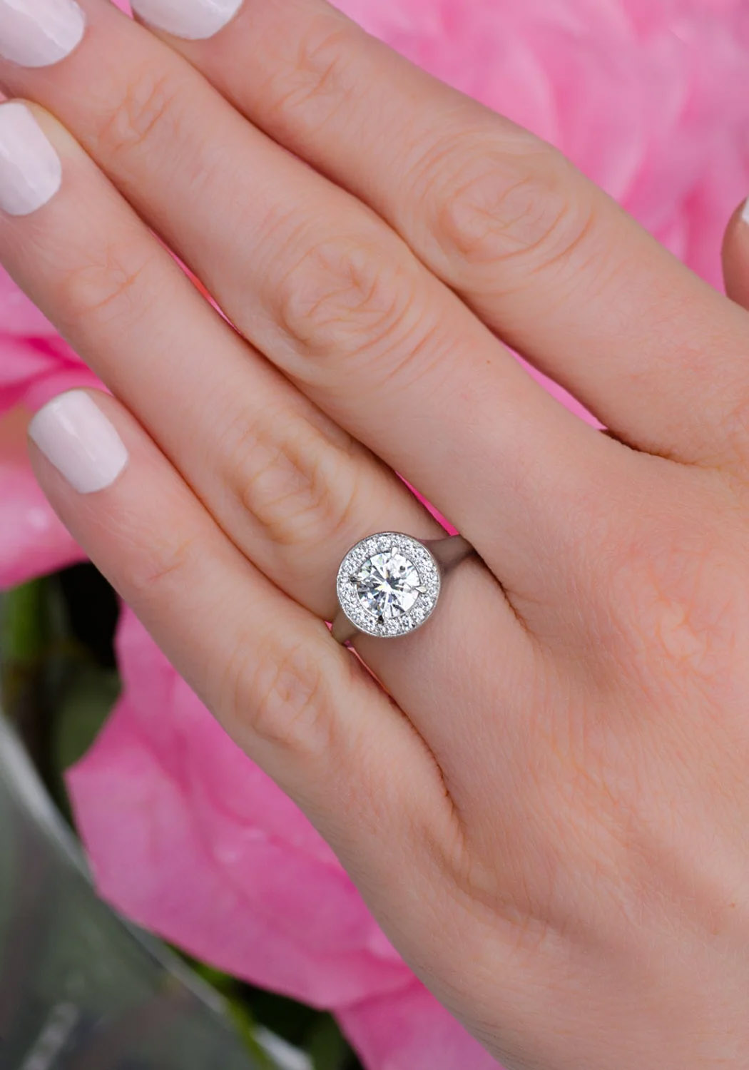 Rahaminov Matte 18KWG Halo Bezel Set Round Diamond Ring on a model's hand in front of a pink bouquet